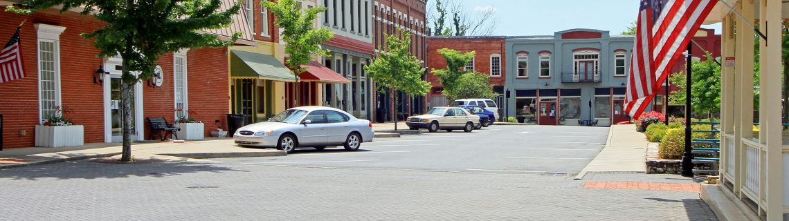 Paved street in a small town with storefronts, parked cars and US flags